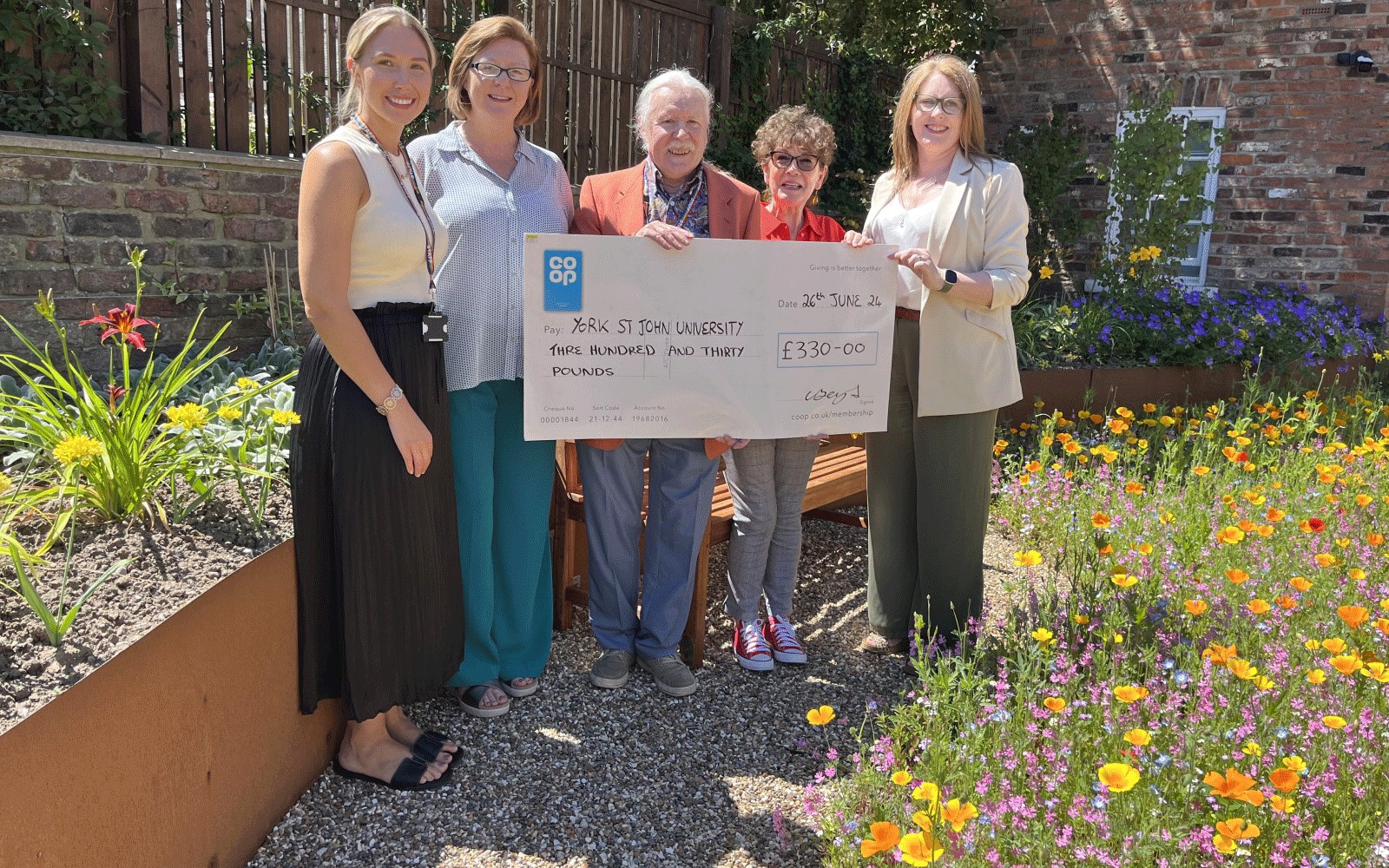 Five people standing in a flower garden holding an oversized cheque