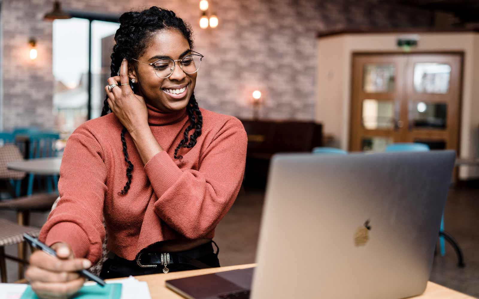 A student sits at a desk with a notepad, looking at a video call on her laptop