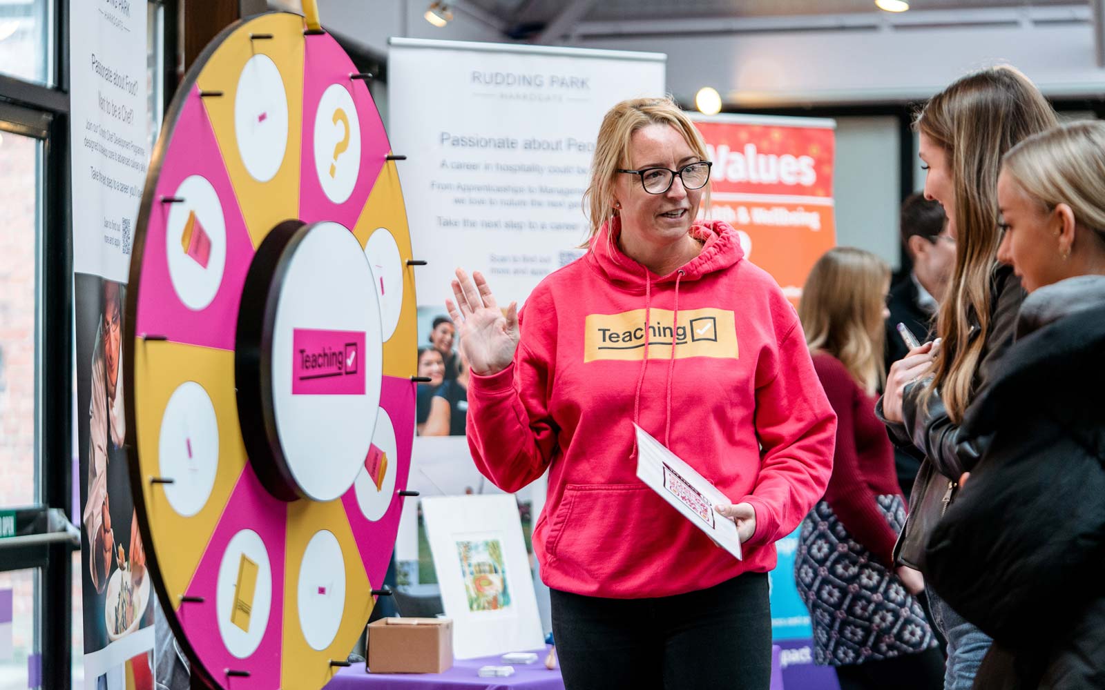 Representative from Get into Teaching with spinning wheel at Careers Fair talking to students.