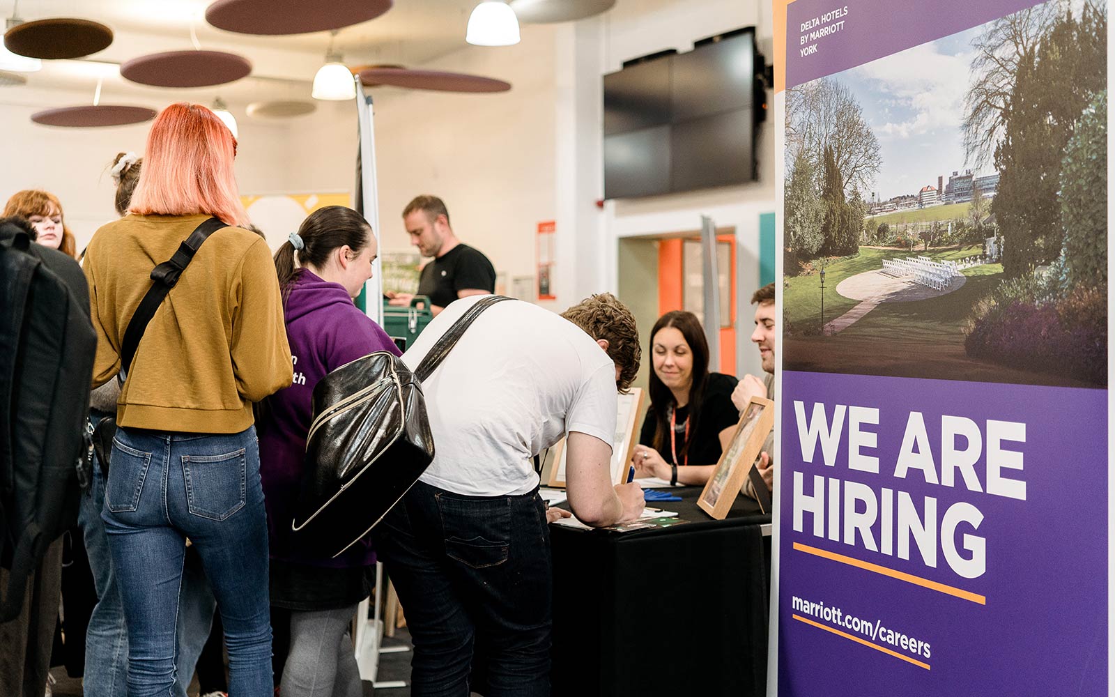 Students signing up at table next to 'We're hiring' sign
