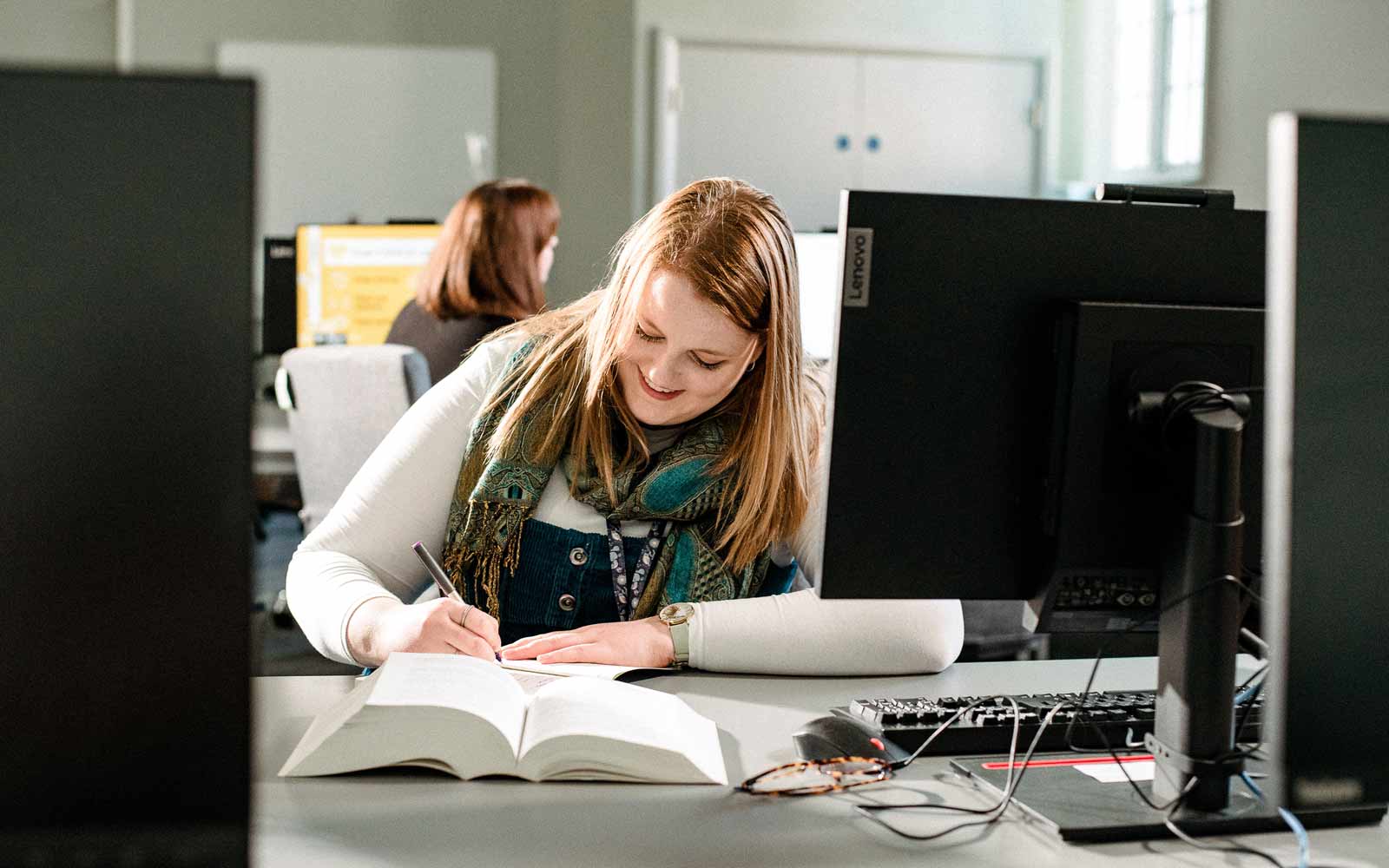 A student making notes and sitting at a desk with a computer, open book, and pair of glasses on it.