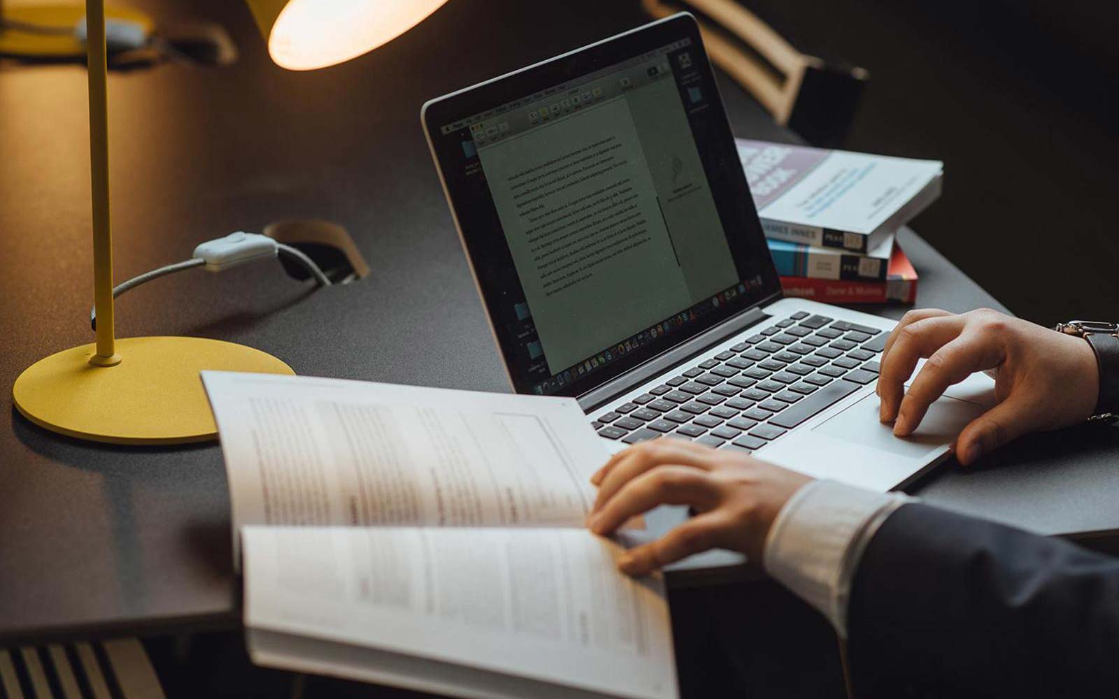 A person working from a on a laptop and reading a book