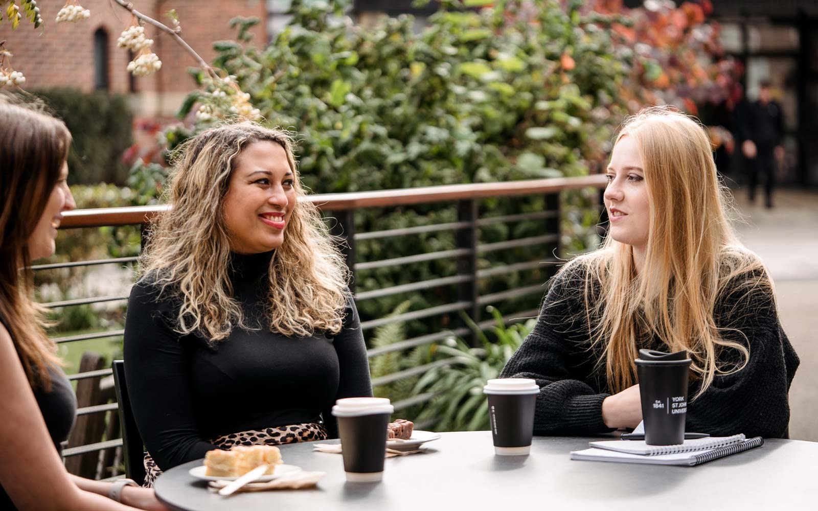 Students sat at table outside on campus chatting and drinking hot drinks