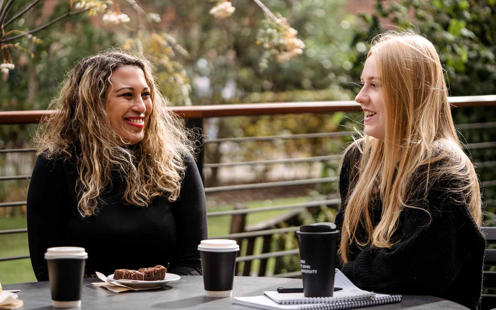 Two staff members sitting at a table outside with coffee cups and a cake.