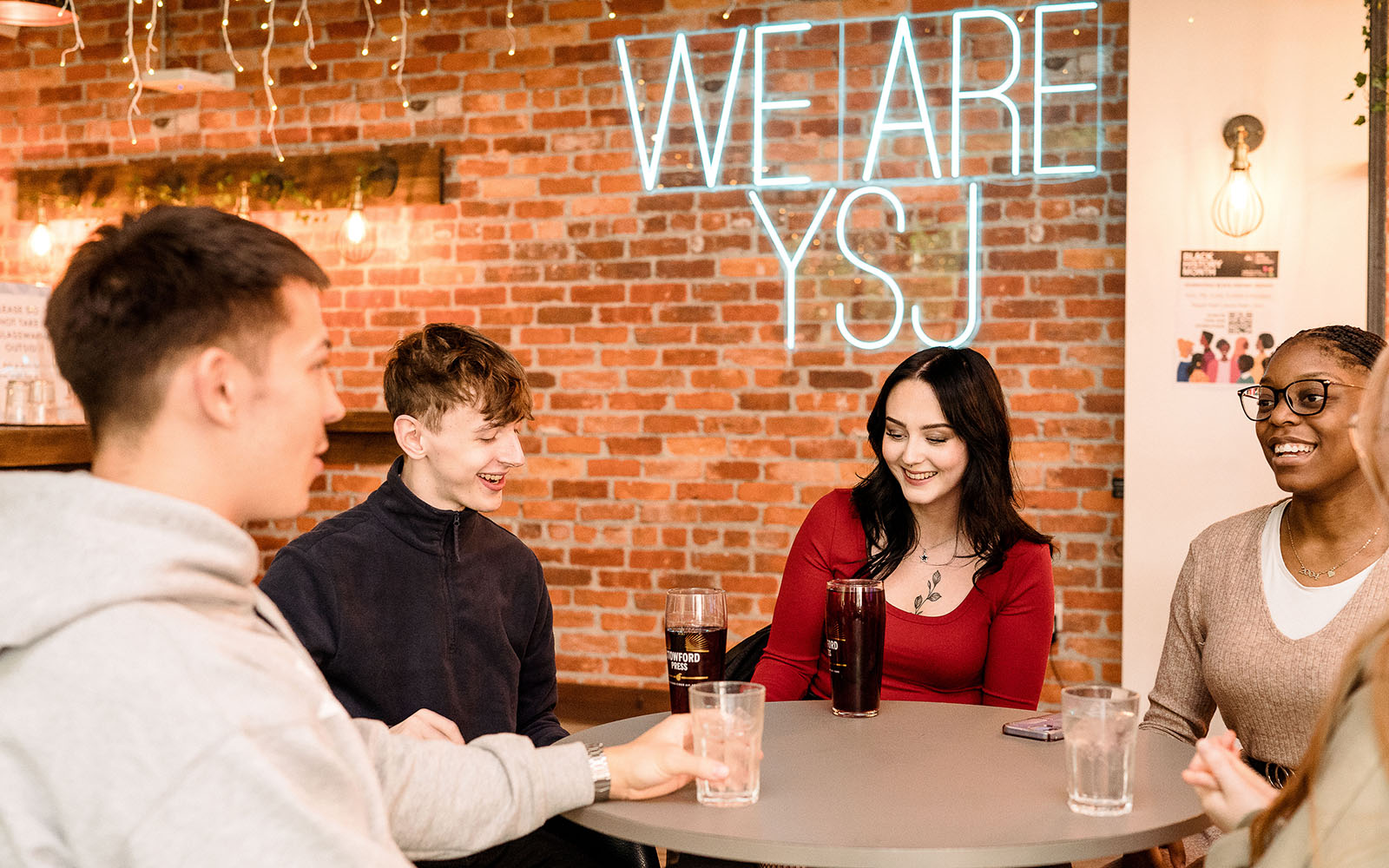Group of students in SU sat round table with drinks in front of We are YSJ sign