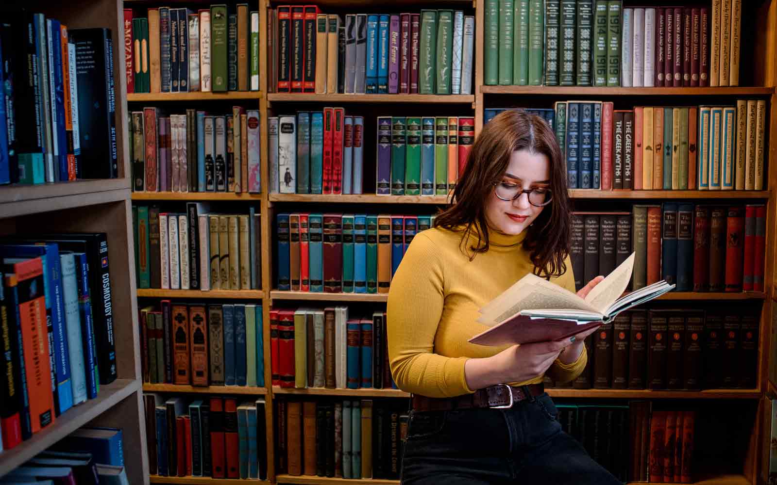 A student holding and looking at an open book and standing near bookshelves.