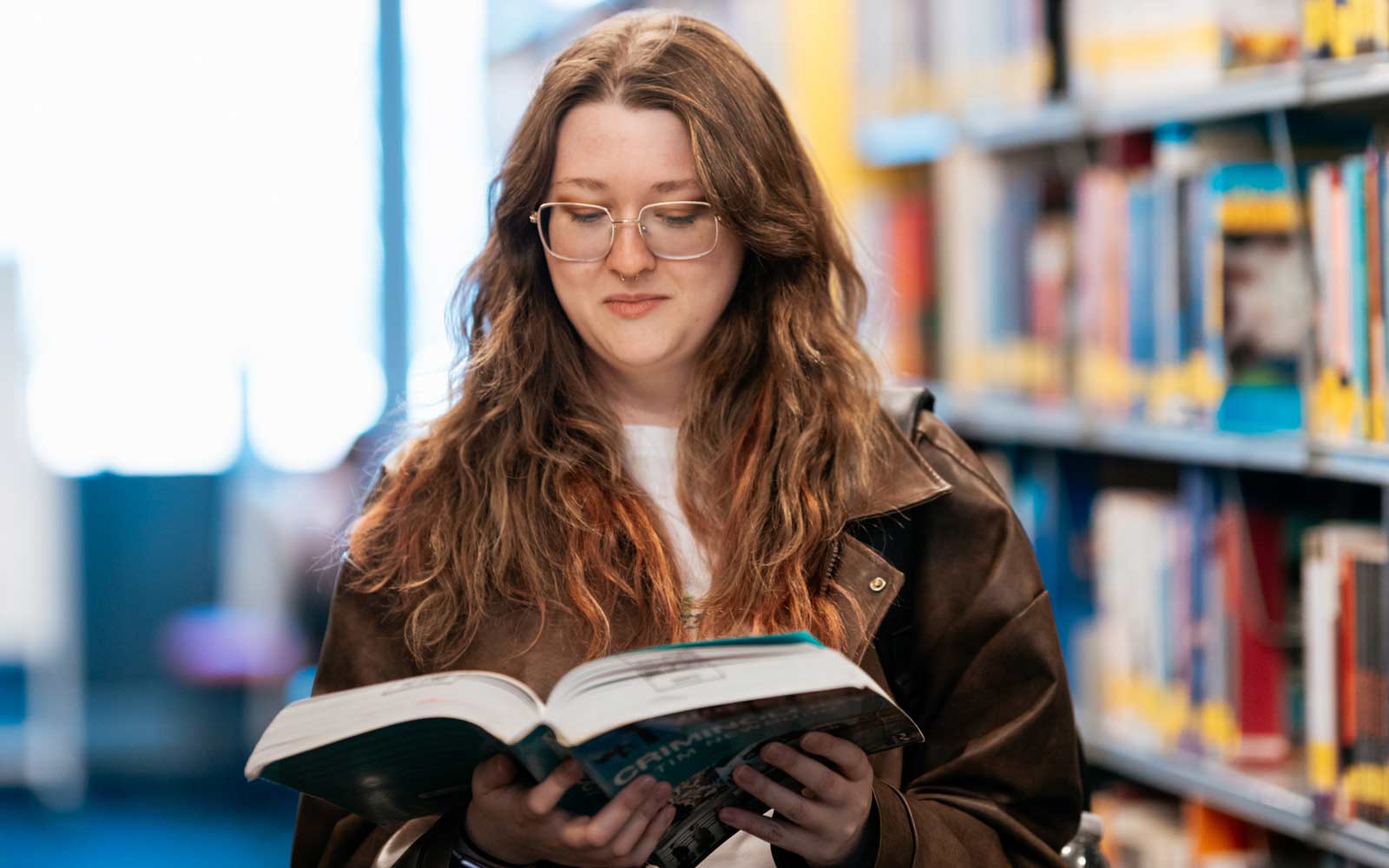 A student standing near the library bookshelves and holding an open book.