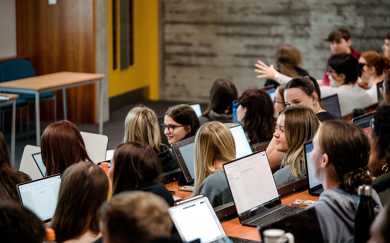 A photo taken over the shoulders of a large group of students sat in a lecture theatre, many with laptops open