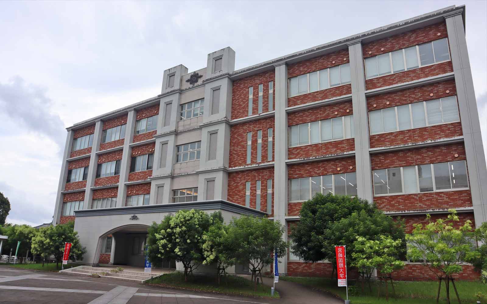 Brick building with clocktower and tree-lined plaza on Miyazaki International College campus.