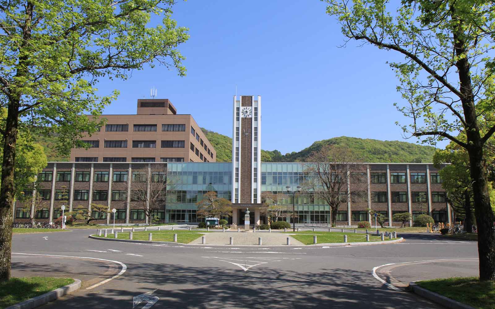 Modern glass-fronted building with clocktower, surrounded by green space and trees with a circular plaza.