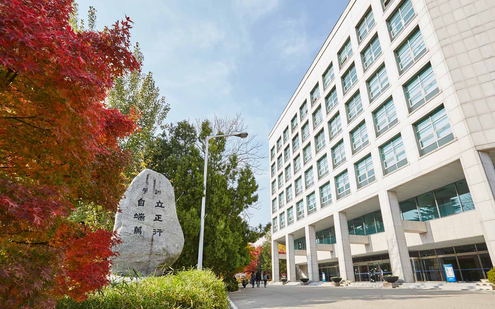 Tall white campus building, with autumn trees and stone Woosong University sign.