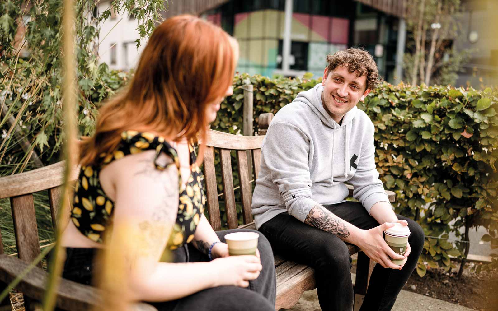 Two people sat on bench in grounds of York St John, drinking coffee and chatting.