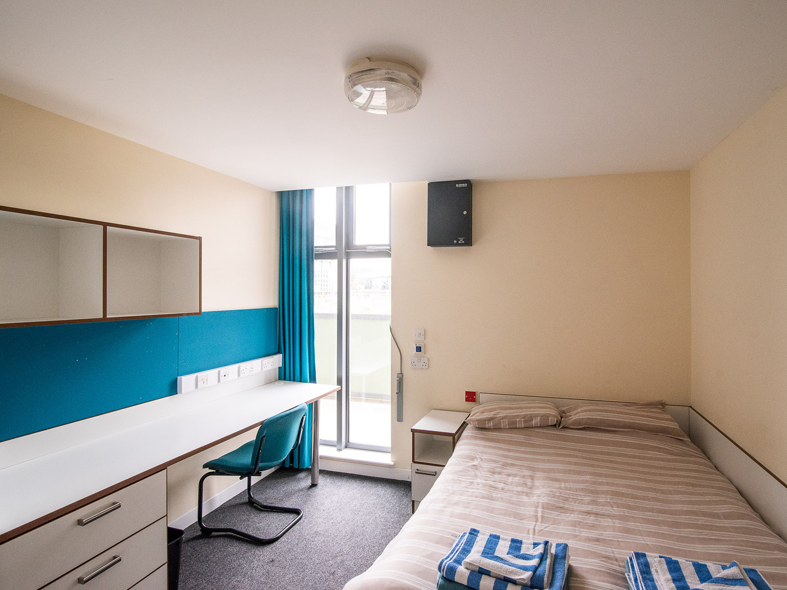 A photograph of accommodation shows a double bed, a bedside table, a floor length window, shelving, and a long desk with a chair and drawers.