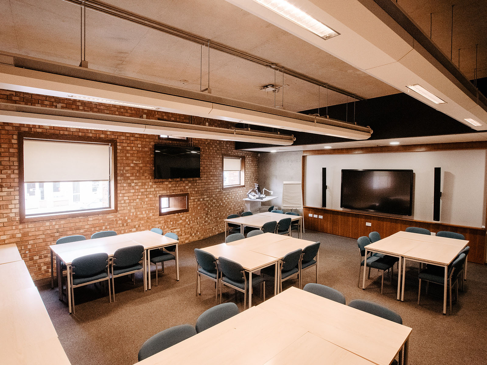 A classroom featuring a lectern, presentation screens, speakers, a flip chart, and reconfigurable desks with space for around 30 chairs