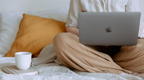A person sits on a bed with a grey laptop on their knees. A while mug is on the bed next to them.