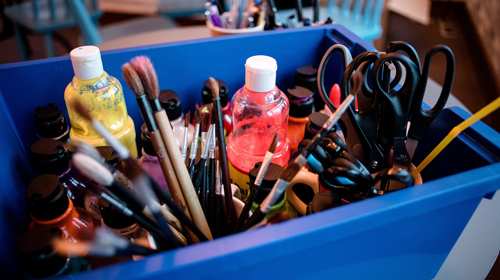 A box of craft materials including paint, brushes and scissors