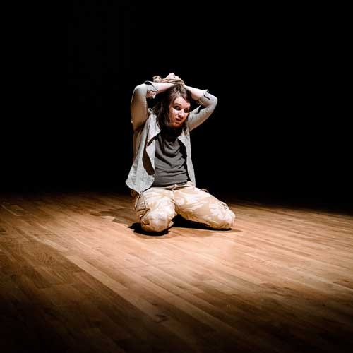Drama student performing. They are knelt on the floor in a spotlight with their hands on their head.