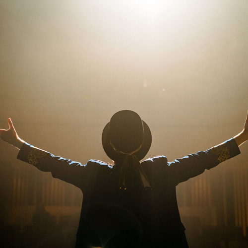 Student from behind on stage wearing top hat with arms stretched out, looking out to audience