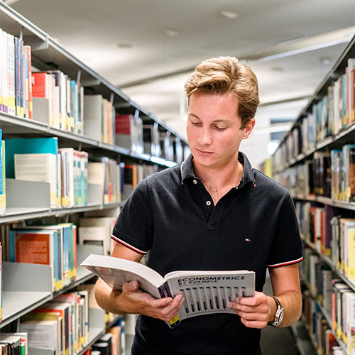 Business student reading Economics book in front of library shelves