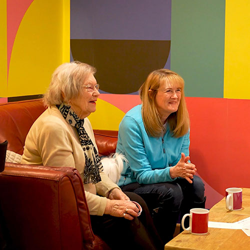Two people sitting on sofa in front of brightly coloured wall